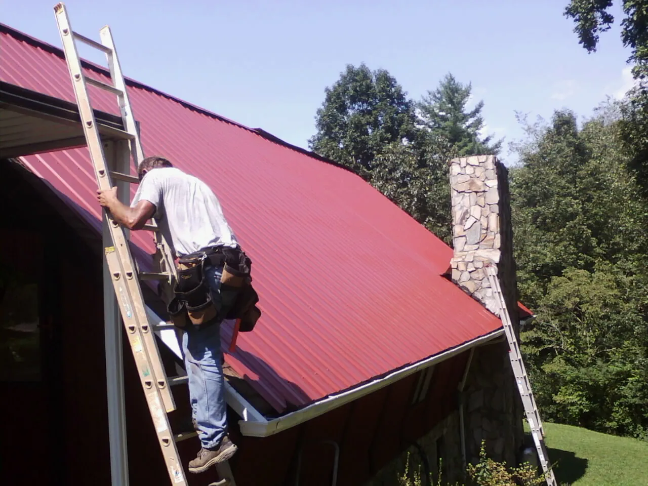 Roofer on a ladder beside a red metal roof and stone chimney — Soffit & Fascia Repair by Roofing Brothers in Pine Plains